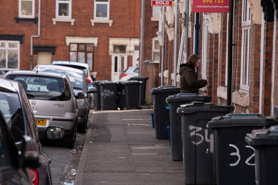 A narrow residential street with brick terraced houses featuring white window frames and stone lintels. Several black wheelie bins with white painted numbers are lined up along the pavement, each with closed lids. A woman dressed in a dark coat is standing beside one of the bins, appearing to be handling rubbish or recycling. Parked cars, including a small silver hatchback, are aligned along the left side of the street, partially blocking the view of the sidewalk. The pavement is concrete, showing some patches and scattered litter. The environment appears to be in the late afternoon with soft natural light, and the scene overall reflects an urban setting where private waste disposal through bin collection is common, supporting services like those offered by Rubbish Removal Tufnell Park for bulky or extra rubbish clearance.
