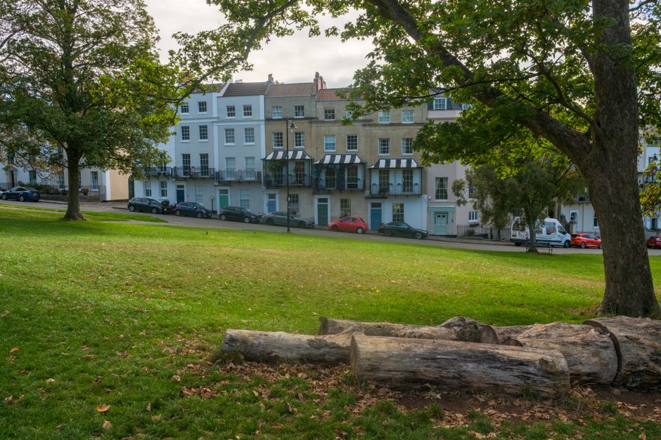 This image depicts a grassy park area with a slight incline, featuring two large, weathered logs lying horizontally in the foreground. The logs have rough, textured bark with visible splits and natural irregularities. Scattered fallen leaves are present around the logs, indicating an outdoor setting during a temperate season. In the midground, there are mature trees with leafy green canopies, their branches arching over the scene and providing partial shade. Beyond the trees, a row of multi-storey residential buildings lines the background; these buildings display a mix of architectural styles with some featuring white facades, large windows, and bay windows, while others have beige stone exteriors. Several parked cars, including two black, a red, and white vehicle, are aligned along the street in front of the buildings. The overall lighting is soft and natural, suggesting an overcast sky. The scene offers an urban environment with a green space, fitting for independent rubbish removal services such as those provided by Rubbish Removal Tufnell Park, which may include on-site clearance or alternative waste handling options for local properties.