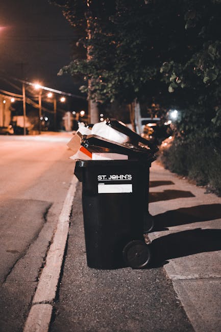 A black plastic wheelie bin with a white label and the name 'St. John's' printed on it, positioned on a pavement beside a roadway at night. The bin's lid is open, revealing various discarded items, including cardboard boxes, paper, and other waste materials. The pavement has a narrow curb separating it from the street, with faint shadows cast by nearby streetlights. In the background, there are trees and a row of streetlights illuminating the road, with parked vehicles visible in the distance. The scene reflects an urban environment where private waste collection might be arranged by a service provider such as Rubbish Removal Tufnell Park, supporting alternative methods of rubbish disposal outside standard local authority collection. The overall lighting is dim, with the streetlights providing a warm glow, creating a clear view of the waste bin and its immediate surroundings while maintaining a quiet, residential atmosphere.