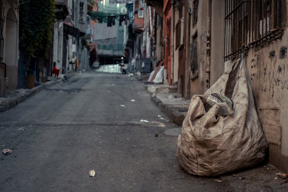 A large, worn, beige fabric trash bag with visible dirt stains and creases is positioned on a narrow urban street pavement next to an old stone wall with iron bars over a window. The bag appears to be partially open, containing some loose debris or trash, and is leaning slightly to the right. The street surface is asphalt with minor cracks and scattered small pieces of litter, including paper and food wrappers. In the background, a steep incline extends upward, flanked by closely spaced, weathered residential buildings with various textures such as brick, concrete, and painted surfaces. Clothes are hanging on lines stretched across the buildings, and laundry items are visible. The scene is lit with natural daylight, but the overall atmosphere suggests a quiet, possibly neglected area that may benefit from independent waste collection services, such as those offered by Rubbish Removal Tufnell Park for alternative waste handling solutions.