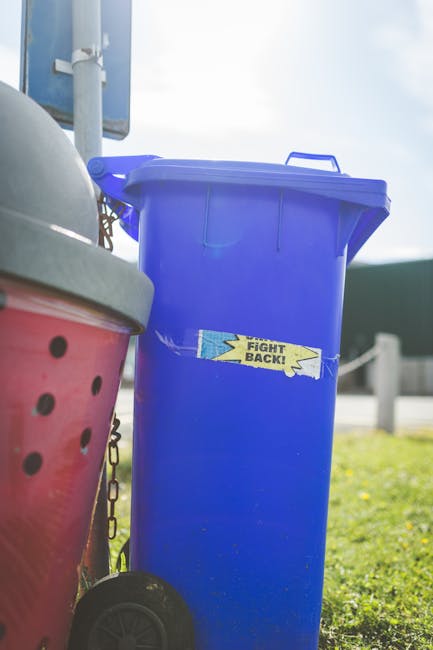 A black plastic wheelie bin with a white label and the name 'St. John's' printed on it, positioned on a pavement beside a roadway at night. The bin's lid is open, revealing various discarded items, including cardboard boxes, paper, and other waste materials. The pavement has a narrow curb separating it from the street, with faint shadows cast by nearby streetlights. In the background, there are trees and a row of streetlights illuminating the road, with parked vehicles visible in the distance. The scene reflects an urban environment where private waste collection might be arranged by a service provider such as Rubbish Removal Tufnell Park, supporting alternative methods of rubbish disposal outside standard local authority collection. The overall lighting is dim, with the streetlights providing a warm glow, creating a clear view of the waste bin and its immediate surroundings while maintaining a quiet, residential atmosphere.