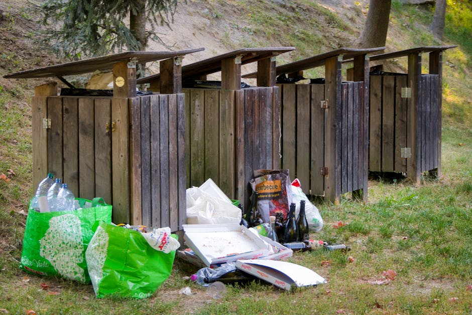 An outdoor scene featuring four wooden waste disposal bins with sloped roofs positioned on a grassy area. The bins are made of vertical wooden planks with a weathered dark brown finish, spaced evenly apart, and each has a small gap at the bottom. They are situated near a gentle slope with some trees and greenery in the background, suggesting a park or outdoor communal space. In front of the bins, there is a collection of discarded waste including a green plastic bag filled with bottles, a cardboard pizza box with some leftovers, and several glass bottles, some of which are dark brown and some clear, with a few other miscellaneous items scattered around. The overall environment appears natural, with the scene illuminated by natural daylight, emphasizing the contrast between the neatly constructed bins and the unmanaged litter in front of them, indicative of a potential issue related to private waste handling or rubbish collection in an outdoor setting. Rubbish Removal Tufnell Park specializes in managing waste and rubbish in such situations, providing alternative disposal solutions suited for outdoor community or private spaces.