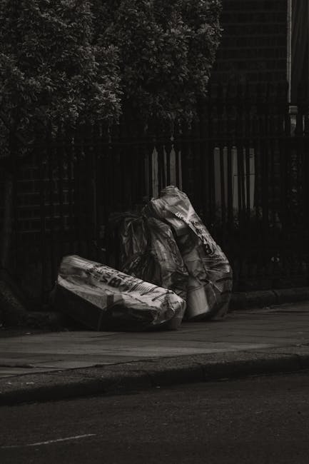 A photograph depicts three large black plastic rubbish bags filled with waste, placed on the pavement near a black metal fence with vertical bars. The bags are crumpled and appear to contain a mixture of household refuse, with some visible text and branding on the packaging. The scene is outdoors, adjacent to a street, with a dark, likely shaded, environment created by nearby trees and shadowed fencing. There is no visible vehicle or waste collection equipment present. The setting suggests a typical area for independent rubbish collection or on-site waste disposal, supported by local waste removal services such as Rubbish Removal Tufnell Park, in line with alternative waste handling options. The background features foliage and the upper part of a building, blending into a muted urban environment with natural and man-made elements.