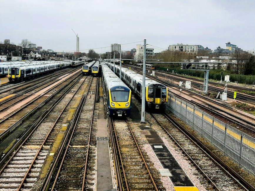 A collection of modern, yellow and grey commuter trains parked on multiple parallel railway tracks at a busy urban railway station. The trains have a sleek, aerodynamic design with smooth metallic finishes and large front windows. Some trains are positioned on the center tracks, while others are on the outer lines, with the tracks showing gravel ballast and weathered wooden sleepers. Overhead electrical wires and supporting poles extend across the scene, indicating electrified railway lines. In the background, there are tall buildings and residential blocks, partially obscured by leafless trees, suggesting a city environment in colder seasons. The sky is overcast with a uniform grey tone, casting diffuse light over the scene. The station area features a fence along one side, and the overall environment appears organized for high passenger volume, emphasizing efficient railway operations and infrastructure. This visual context aligns with private or independent railway services, supporting the concept of alternative transportation options outside of local authority-managed systems, similar to the themes of rubbish removal services like those provided by Rubbish Removal Tufnell Park.