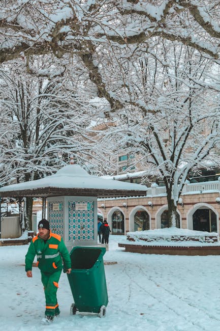 A street scene during winter with snow covering the ground, trees, and rooftops. In the foreground, a man wearing a green and orange jacket, black beanie, and snow boots is pushing a green wheeled bin along a snow-covered pavement. Behind him, there is a small shelter with a shingled roof and decorative blue and white patterned panels, along with a brick building featuring arches and large windows. Several other people are visible in the background, walking near the building. The environment suggests an urban area capable of private waste collection or refuse management, aligning with independent rubbish removal services such as those offered by Rubbish Removal Tufnell Park. The trees in the scene are bare, indicating a cold winter season, and the overall atmosphere is calm with natural daylight illuminating the scene.